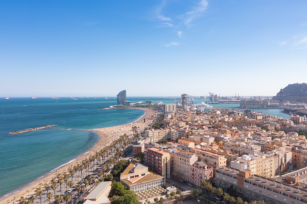 Luftbild der Barceloneta: links Strand und Uferpromenade, rechts die dicht bebauten Blöcke des Viertels, dahinter der Hafen