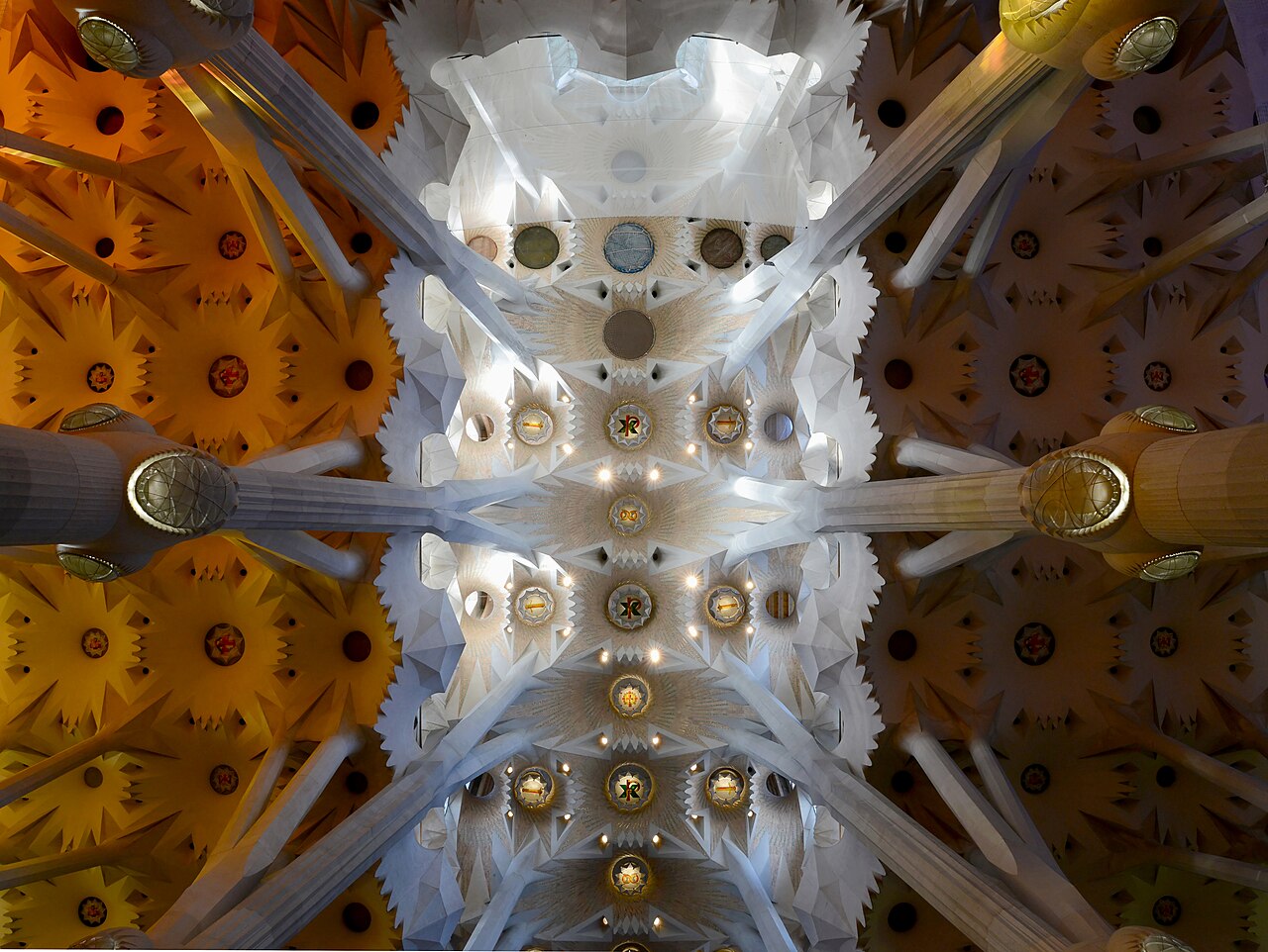 Blick senkrecht nach oben auf die Decke des Kirchenschiffs der Sagrada Família mit den baumartigen Säulen und farbigem Licht
